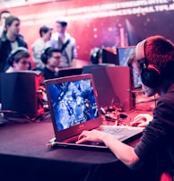 Several people are seated and focused on computer screens, wearing headsets, in an environment that appears to be a gaming tournament or esports event. The room is dimly lit with neon lighting, highlighting screens and creating an intense atmosphere.