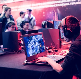 Several people are seated and focused on computer screens, wearing headsets, in an environment that appears to be a gaming tournament or esports event. The room is dimly lit with neon lighting, highlighting screens and creating an intense atmosphere.