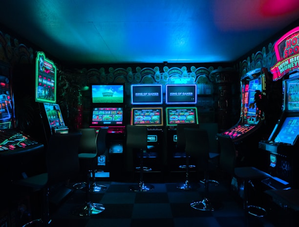 Dimly lit gaming arcade featuring a variety of brightly illuminated slot machines and video gaming screens. Neon signs and colorful displays create a vibrant atmosphere. The arrangement of gaming machines suggests an inviting space for entertainment, with chairs positioned for players.