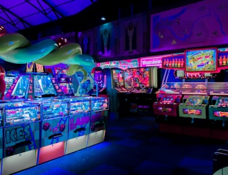 An indoor arcade filled with vibrant gaming machines lit with neon blue and pink lights. The scene includes various arcade games with flashing screens and colorful displays, and a large overhead structure resembling a green snake. The background shows illuminated signs and themed decor, contributing to an immersive entertainment atmosphere.