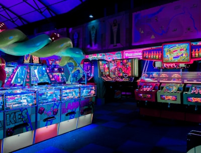 An indoor arcade filled with vibrant gaming machines lit with neon blue and pink lights. The scene includes various arcade games with flashing screens and colorful displays, and a large overhead structure resembling a green snake. The background shows illuminated signs and themed decor, contributing to an immersive entertainment atmosphere.