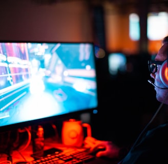 A person wearing headphones is sitting in front of a computer monitor. The screen displays a vibrant, illuminated video game scene, while the person appears focused on gaming. The ambient lighting is dark with red and blue tones, and a backlight from the monitor highlights the headphones.