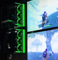 Two gaming monitors display an action scene from a video game featuring a character with purple hair in a lush outdoor setting. The gaming PCs are mounted on the wall with glowing green LED lights, and the room is dimly lit to emphasize the gaming setup.