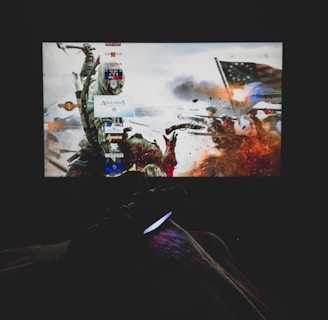 A dimly lit room with a television screen displaying a dynamic, action-packed video game interface featuring a historical battle scene. The foreground shows the silhouette of a gaming console controller held by a person.
