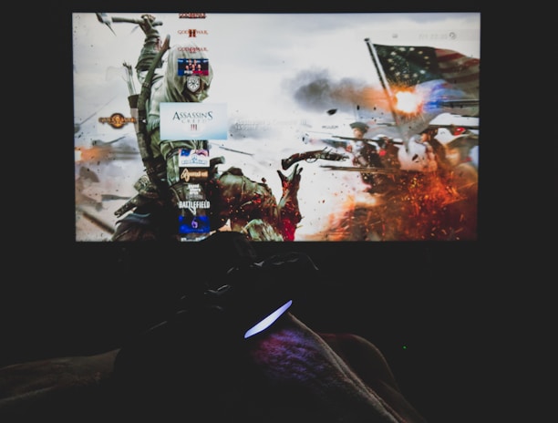 A dimly lit room with a television screen displaying a dynamic, action-packed video game interface featuring a historical battle scene. The foreground shows the silhouette of a gaming console controller held by a person.