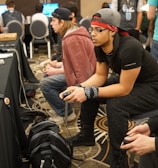 Several people are sitting in a room with gaming consoles and monitors, focusing intently on their screens. The setting appears to be a casual gaming event, with participants holding controllers while playing video games. The room has a casual atmosphere with chairs and tables set up for gaming.