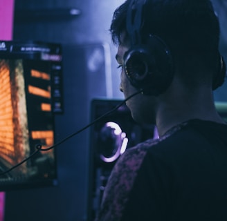 A person wearing headphones is engaged in gaming on a computer, with a large screen displaying a video game scene, showing a wooden building and a path.