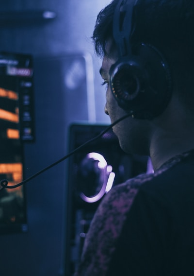 A person wearing headphones is engaged in gaming on a computer, with a large screen displaying a video game scene, showing a wooden building and a path.