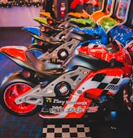 A row of arcade motorcycle racing games is lined up, featuring brightly colored machines with detailed graphics and logos. A person in the background stands near decorated countertops and a wall-mounted screen. The environment has a lively and playful ambiance, with vibrant colors and patterns.