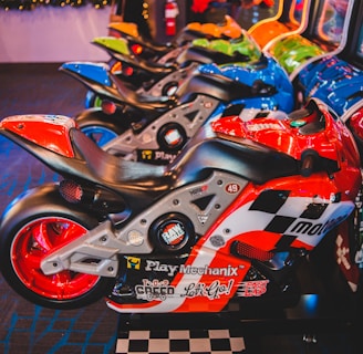 A row of arcade motorcycle racing games is lined up, featuring brightly colored machines with detailed graphics and logos. A person in the background stands near decorated countertops and a wall-mounted screen. The environment has a lively and playful ambiance, with vibrant colors and patterns.