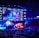 A large esports arena featuring a massive screen displaying a video game, surrounded by sponsor logos and team names. The auditorium is filled with spectators watching the gaming event. Neon blue and purple lights illuminate the stage, creating a vibrant atmosphere.