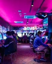A vibrant arcade with rows of gaming machines adorned with colorful screens. Several people are immersed in playing games, seated on stools in a dimly lit room accented by an intense pink ceiling light. The atmosphere is lively and engaging.