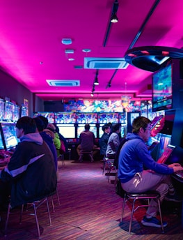 A vibrant arcade with rows of gaming machines adorned with colorful screens. Several people are immersed in playing games, seated on stools in a dimly lit room accented by an intense pink ceiling light. The atmosphere is lively and engaging.