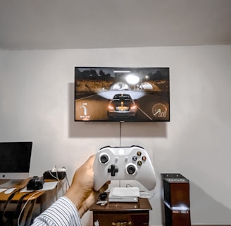 A person is holding a gaming controller in front of a flat-screen television displaying a racing video game. The room has a computer setup on a wooden desk with cables, a desktop computer monitor, and other accessories. The focus is on the interaction between the person and the gaming environment.