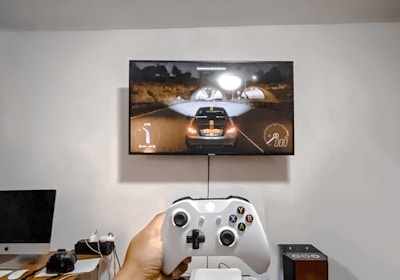 A person is holding a gaming controller in front of a flat-screen television displaying a racing video game. The room has a computer setup on a wooden desk with cables, a desktop computer monitor, and other accessories. The focus is on the interaction between the person and the gaming environment.