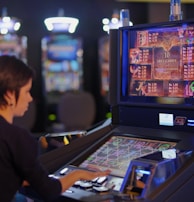 A person is seated at a slot machine console in a casino, focusing intently on the game displayed on the screen. The background shows several other illuminated gaming machines, adding a vibrant and colorful atmosphere to the scene.