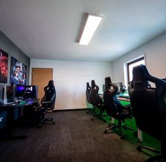 A well-lit gaming room features multiple gaming stations with high-backed chairs and monitors. The room has a minimalist setup with green and black color tones. Large posters adorn the walls, and the room appears clean and organized.