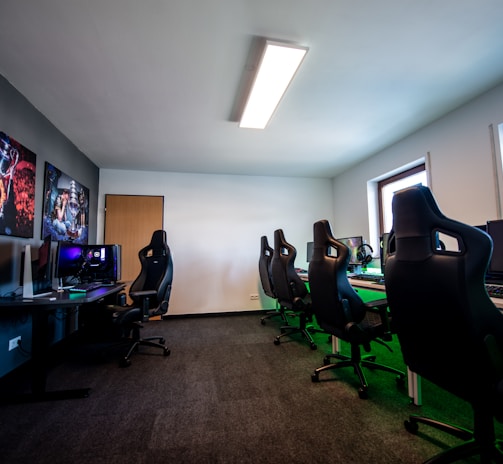A well-lit gaming room features multiple gaming stations with high-backed chairs and monitors. The room has a minimalist setup with green and black color tones. Large posters adorn the walls, and the room appears clean and organized.
