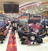 A bustling arcade filled with people playing various gaming machines. Brightly colored displays and animated characters are prominent on posters and screens. The atmosphere is lively with patrons seated in rows, focusing on the games. Overhead signage and electronic scoreboards are visible, contributing to the busy and vibrant environment.