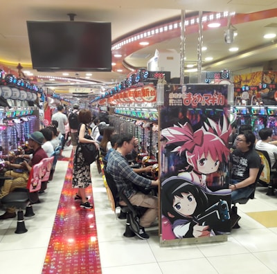 A bustling arcade filled with people playing various gaming machines. Brightly colored displays and animated characters are prominent on posters and screens. The atmosphere is lively with patrons seated in rows, focusing on the games. Overhead signage and electronic scoreboards are visible, contributing to the busy and vibrant environment.