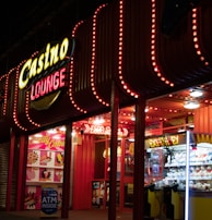 A brightly lit casino lounge entrance adorned with neon lights. The exterior features colorful signage including the words 'Casino' and 'Lounge' in vivid colors, with additional lights framing the building. Inside, a glimpse of a claw machine filled with plush toys and arcade games is visible. The area exudes an inviting yet bustling atmosphere typical of entertainment venues.
