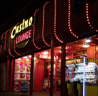 A brightly lit casino lounge entrance adorned with neon lights. The exterior features colorful signage including the words 'Casino' and 'Lounge' in vivid colors, with additional lights framing the building. Inside, a glimpse of a claw machine filled with plush toys and arcade games is visible. The area exudes an inviting yet bustling atmosphere typical of entertainment venues.