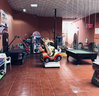 An indoor arcade area featuring various gaming machines, including an air hockey table and a ride-on toy car. The room has a tiled floor and several colorful gaming devices scattered around. Chairs and arcade signage are visible, creating a playful and entertainment-focused atmosphere.