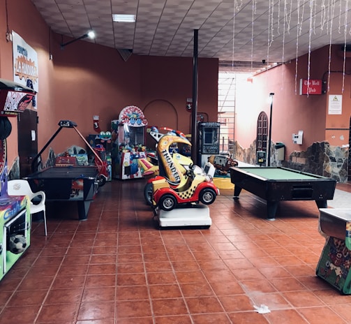 An indoor arcade area featuring various gaming machines, including an air hockey table and a ride-on toy car. The room has a tiled floor and several colorful gaming devices scattered around. Chairs and arcade signage are visible, creating a playful and entertainment-focused atmosphere.
