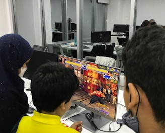 A group of children is gathered around a desktop computer, engaged in playing a colorful, action-packed arcade game displayed on the monitor. The setting appears to be a modern, well-lit office or gaming space with large glass windows, showing multiple computer workstations in the background.
