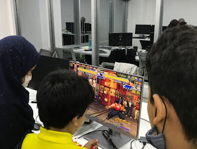 A group of children is gathered around a desktop computer, engaged in playing a colorful, action-packed arcade game displayed on the monitor. The setting appears to be a modern, well-lit office or gaming space with large glass windows, showing multiple computer workstations in the background.