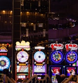 A lively casino scene featuring a row of bright, colorful slot machines with themes like Monopoly, Michael Jackson, and Lord of the Rings. The gaming machines display a variety of flashing lights and digital screens, creating an energetic and exciting atmosphere. Reflections of the lights are visible in glossy surfaces above the machines.