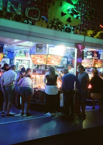 A group of people standing in front of an illuminated amusement arcade, engaging with various gaming machines. The environment is bustling with activity, and colorful lights create a vibrant atmosphere. The ceiling is decorated with large, playful images of dice and other game-related graphics, enhancing the lively mood.