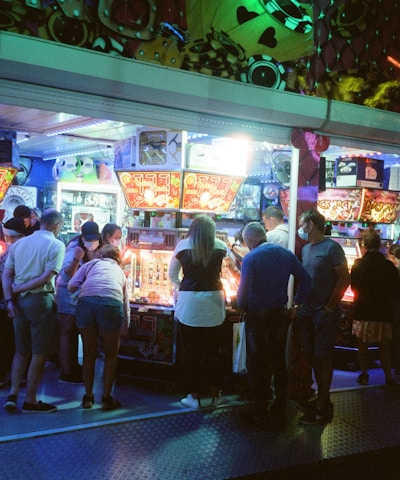 A group of people standing in front of an illuminated amusement arcade, engaging with various gaming machines. The environment is bustling with activity, and colorful lights create a vibrant atmosphere. The ceiling is decorated with large, playful images of dice and other game-related graphics, enhancing the lively mood.