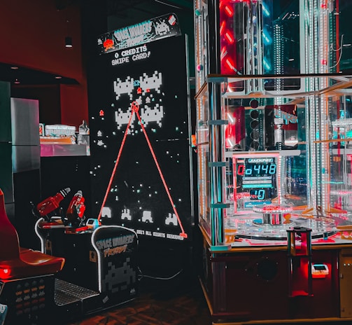 An arcade setting featuring a classic Space Invaders game setup and a modern gaming machine with seats. Bright neon lights illuminate the area, with scores displayed on digital screens. A bright and visually engaging environment full of electronic components.