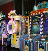 A row of brightly colored arcade game machines is arranged in a line against the wall. Each machine features vibrant lights and designs, with themes ranging from claw games to game show-inspired setups. The area has a playful and inviting atmosphere, suitable for entertainment or amusement centers.