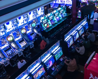 A bustling arcade filled with rows of gaming machines, each with vibrant displays and controls. Several people are seated at these machines, deeply engaged in playing games. Bright fluorescent lights cast a blue and neon glow across the room, enhancing the lively and energetic atmosphere.