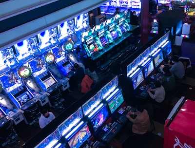 A bustling arcade filled with rows of gaming machines, each with vibrant displays and controls. Several people are seated at these machines, deeply engaged in playing games. Bright fluorescent lights cast a blue and neon glow across the room, enhancing the lively and energetic atmosphere.