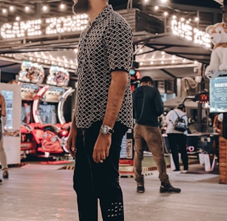 A person stands in a lively arcade setting at night, surrounded by string lights and various arcade game machines. The scene is bustling with activity as other people are visible in the background, engaging with the games or walking by. The signage reads 'Game Zone', indicating the entertainment venue.