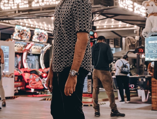 A person stands in a lively arcade setting at night, surrounded by string lights and various arcade game machines. The scene is bustling with activity as other people are visible in the background, engaging with the games or walking by. The signage reads 'Game Zone', indicating the entertainment venue.