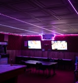 A dimly lit room with vibrant purple LED strip lights on the ceiling, featuring multiple gaming stations. There are two large screens showing video games, along with various arcade machines, including a Nintendo arcade cabinet. An air hockey table is positioned in the foreground.