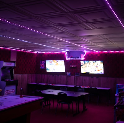 A dimly lit room with vibrant purple LED strip lights on the ceiling, featuring multiple gaming stations. There are two large screens showing video games, along with various arcade machines, including a Nintendo arcade cabinet. An air hockey table is positioned in the foreground.