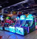 A brightly lit arcade filled with colorful gaming machines sits under ceiling lights. Neon lights illuminate various game stations, creating a playful and vibrant environment. A few red chairs are scattered around the reflective, tiled floor, and a person is observing one of the machines. The atmosphere is lively and cheerful, featuring games with cartoonish designs and engaging themes.