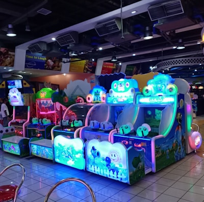 A brightly lit arcade filled with colorful gaming machines sits under ceiling lights. Neon lights illuminate various game stations, creating a playful and vibrant environment. A few red chairs are scattered around the reflective, tiled floor, and a person is observing one of the machines. The atmosphere is lively and cheerful, featuring games with cartoonish designs and engaging themes.