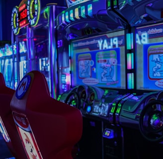 A vibrant arcade environment with several illuminated gaming machines. The image features arcade racing games with visible steering wheels, colorful control panels, and screens displaying game instructions. Bright LED lights in various colors illuminate the machines and surrounding area.