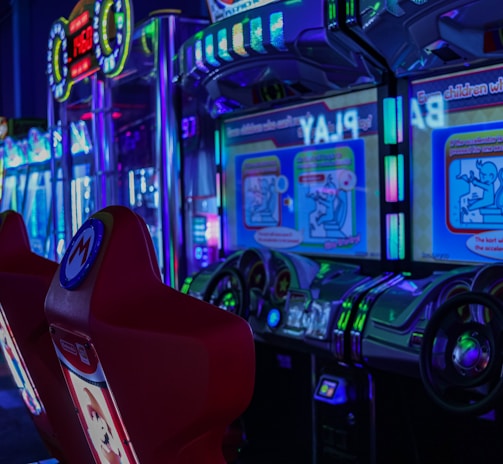 A vibrant arcade environment with several illuminated gaming machines. The image features arcade racing games with visible steering wheels, colorful control panels, and screens displaying game instructions. Bright LED lights in various colors illuminate the machines and surrounding area.