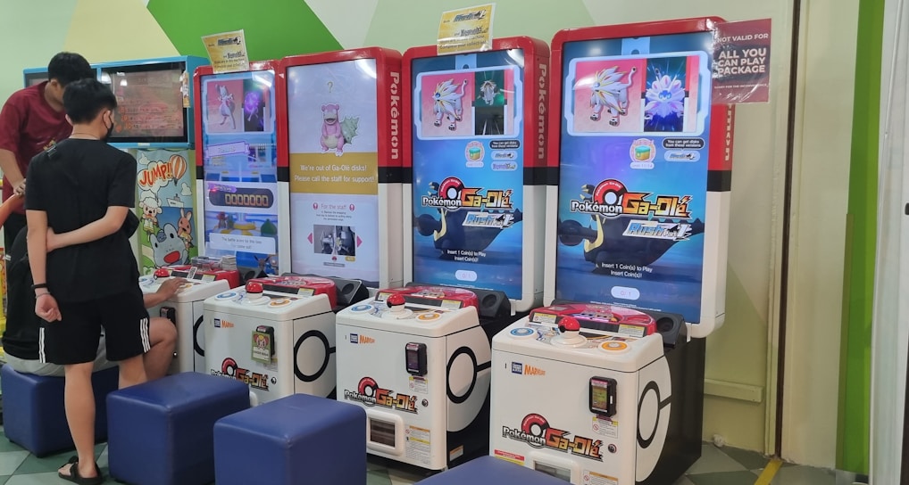 Several arcade gaming machines with multiple screens displaying Pokémon-themed content are positioned in a colorful arcade setting. There are people interacting with the machines; some are seated, and others are standing. The area features different gaming options, including a jumping game with bright, inviting graphics.