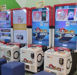 Several arcade gaming machines with multiple screens displaying Pokémon-themed content are positioned in a colorful arcade setting. There are people interacting with the machines; some are seated, and others are standing. The area features different gaming options, including a jumping game with bright, inviting graphics.